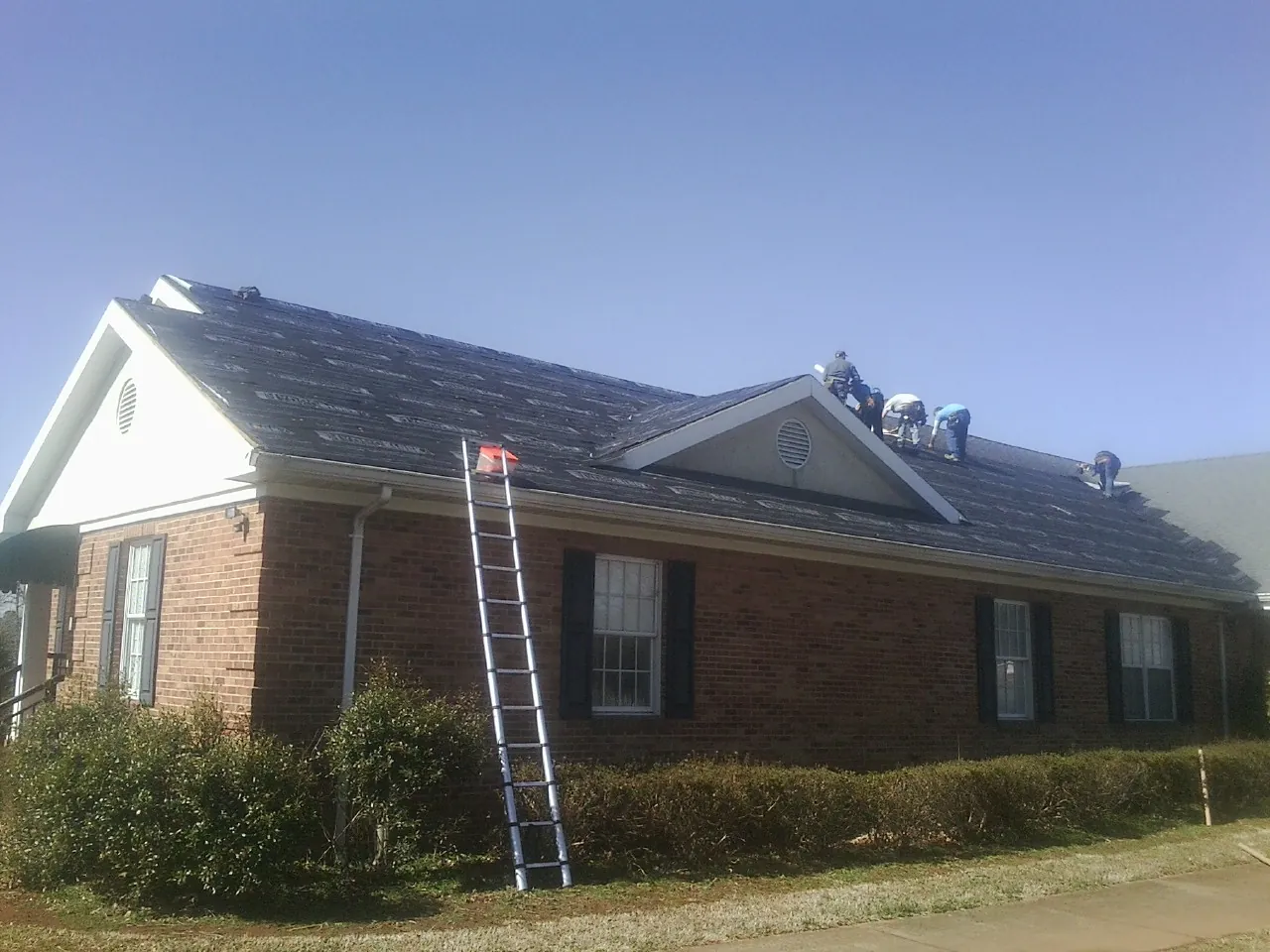 Crew tearing shingles off a brick ranch roof with a ladder set — Metal Roof Repair work by our crew in East Andover