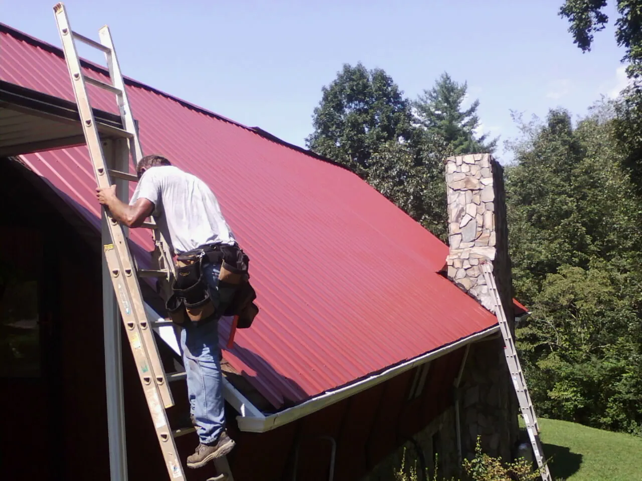 Roofer on a ladder beside a red metal roof and stone chimney — Soffit & Fascia Repair by Roofing Brothers in East Andover